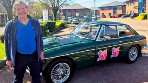 PHA UK Dave Whittington wearing a blue t-shirt and cardigan, standing near the bonnet of his dark green 57-year-old MGC classic car.
