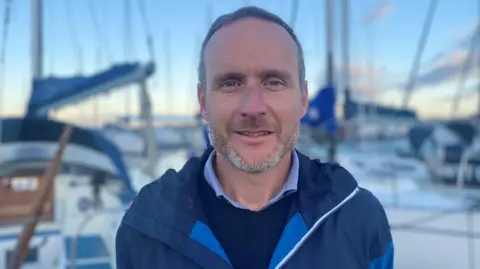 Stuart Fox is at Nairn harbour and there are boats and masts behind him. Stuart has short fair hair and designer stubble. He is wearing a blue jacket over a blue jumper and shirt.