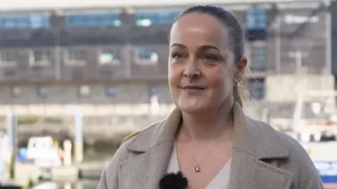 A woman looking towards the camera. She is wearing a light brown coat and has boats and water behind her.