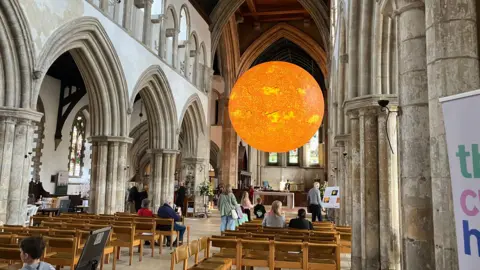 The inside of a tall church has a row of wooden seats and a large bold orange Sun sculpture suspended from the ceiling. Some people are sitting down and others are standing around, looking up at the sculpture.