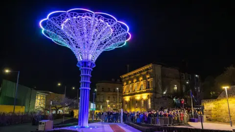 Barnsley Council A towering metal structure has ornate filigree in the shape of a rose and is lit up with blue lights in the night sky. A crown of people are looking up at it