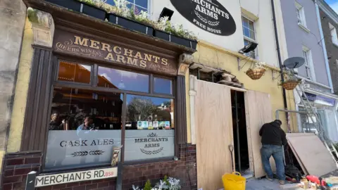An old fashioned pub with a brown tiled exterior and signage reading "The Merchant's Arms". A man is in the process of boarding up a hole in the facade. 