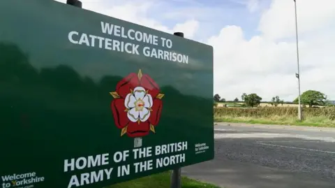 A green sign by a road reading: "Welcome to Catterick Garrison, home of the British Army in the North."