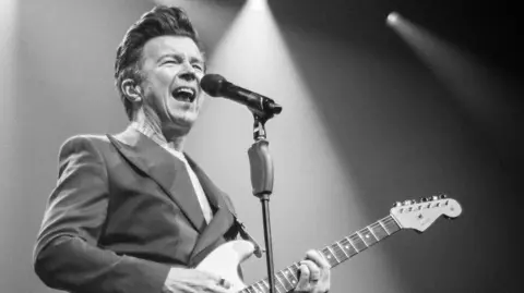 Cuffe & Taylor A black and white photo of a man with brown hair in a quiff style, wearing a suit and holding a guitar. He is mid-song as he sings into a microphone with lighting seen behind him.