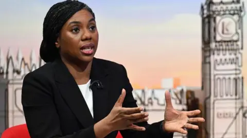 Kemi Badenoch sitting in a red chair is looking at the camera and wearing a white blouse and black suit jacket.