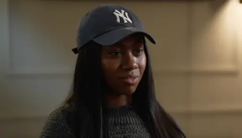 A young black woman in a New York Yankees baseball hat and grey jumper looks to her left, apparently being interviewed