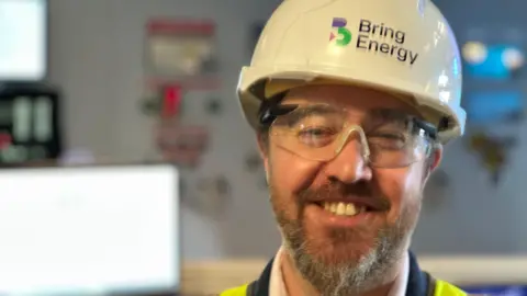 A man wearing a white hard hat and safety goggle. The hat has black writing with the words Bring Energy on it. The man has a stubbly beard. He is smiling for the photograph and is standing indoors.