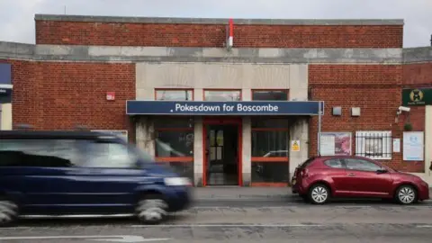 Pokesdown Station is a small red-brick building with a blue name sign above some glass doors. It is on a main road, and cars are driving past it.