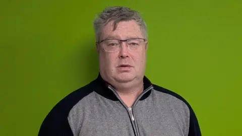 Gavin Harrower Gavin has short grey hair and glasses. He is wearing a grey top with a zip.