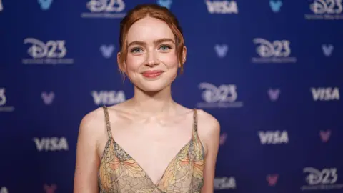 Sadie Sink smiling for the camera, she is wearing a strappy top that has a pattern on it that looks like stained-glass windows. She has auburn hair tied up and behind her is a blue advertising board for Disney's D23 fan event.