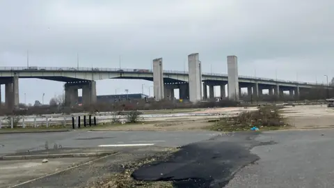 Large bridge in distance vast expanse of unused car park and unkempt weeds in foreground 