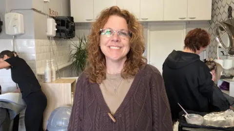 A woman standing in a hairdressers, wearing a brown jumper and a light brown top.