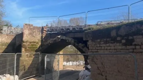 Closed bridge, with barriers on top of it and also near it close to the bottom of the bridge by the river.