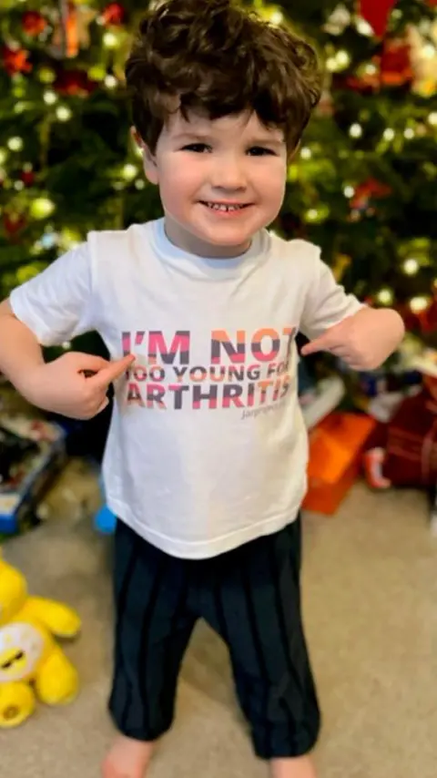 Dean Ranyard Boy smiling with brown hair. He has a white top with red writing that reads "I'm not too young for arthritis". He has dark trousers and bare feet. He has a yellow toy bear near him and a Christmas tree with presents. He is pointing at the writing on his top.