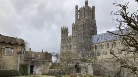 Orla Moore/BBC Ely Cathedral from its south side, showing the west tower and part of the nave against a grey wintery sky. It is shot from a walled garden. 