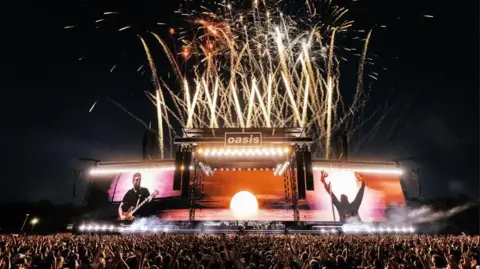 Big Brother Recordings Colourful fireworks light up the sky behind the stage, with large screens showing Noel Gallagher playing guitar and a silhouetted Liam Gallagher raising his arms, holding a tambourine. Many in the crowd are raising their hands in the air.