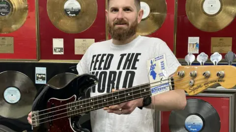 PA Media Auctioneer Luke Hobbs from Gardiner Houlgate stands facing the camera holding a black bass guitar which was played by Chris Cross at Live Aid. Mr Hobbs has a beard and is wearing a white T-shirt with "Feed the World" in black letters on it