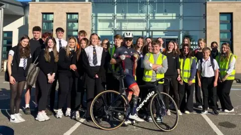 Fin Graham on his bike, holding his silver and gold medals and surrounded by school pupils