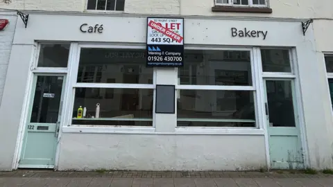 Francesca Griffiths The outside of a shop building on a high street with "Cafe" and "Bakery" written over the windows, either side of a "to let" board which is a red "under offer" sticker across it. The shop has two light green doors with windows at either end, divided by two large glass windows.