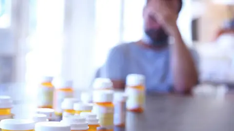 Getty Images A blurred image of a man with a light coloured t-shirt sitting at his table with his head in his hands. In front of him are orange bottles of pills with white lids sitting on the table.