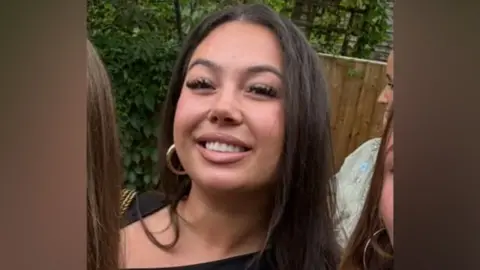 A close-up of a smiling young woman with long dark hair and gold hoop earrings, outdoors against a backdrop of green foliage and a wooden fence.
