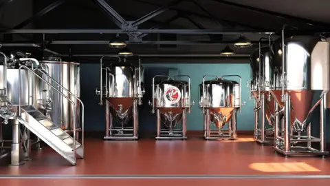 Rows of large silver containers on silver legs in a large room with a brown floor and green walls