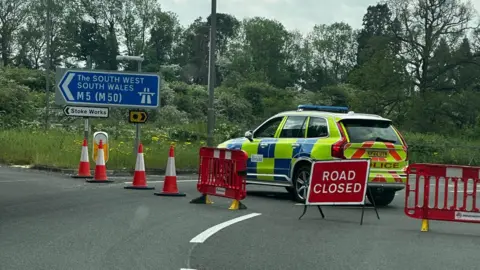 Tracey Ball A high viz yellow and blue 4x4 police car is parked across the road. In front of the police vehicle are orange traffic cones plastic orange barriers. There is also a red square shaped sign that reads "road closed" in white writing.