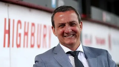 Head and shoulders shot of Andy Pilley sitting in a football stand smiling. He has short dark hair and is wearing a white shirt, black tie and grey suit jacket