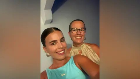 Chloe McGinty Two women are smiling at the camera. They both have dark hair which has been tied back. 