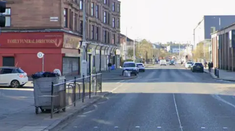 A long road of red sandstone tenements and shops in Glasgow. There are parked cars.
