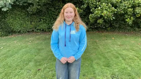 A teenage girl with long wavy ginger hair wearing a blue hoodie and blue denim jeans smiling at the camera and stood in front of a set of shrubs in a garden.