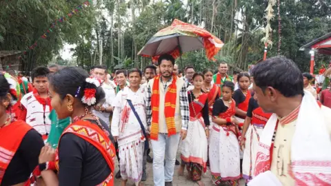 Aditya Sharma Pranab Doley stands under a traditional umbrella, wearing off-white pants, a check shirt with jacket and a bright orange scarf. He is sorrounded by men and women, all dressed in traditional Assamese attire