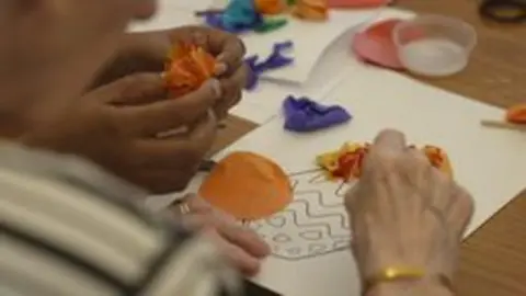 Close-up of two pairs of hands working on a collage - one pair is that of an old person, the other of a younger care worker. This shot was taken in June 2025.