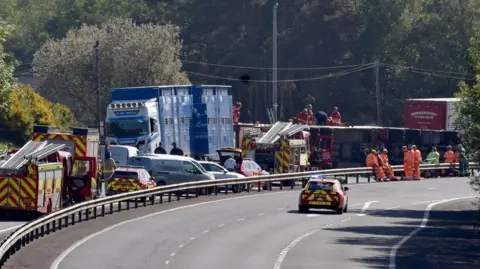 Pacemaker Emergency services attend the scene of a crash on the M1 motorway. An overturned lorry can be seen in the distance with a number of fire appliances parked nearby. Another blue livestock lorry is parked close by the overturned lorry.