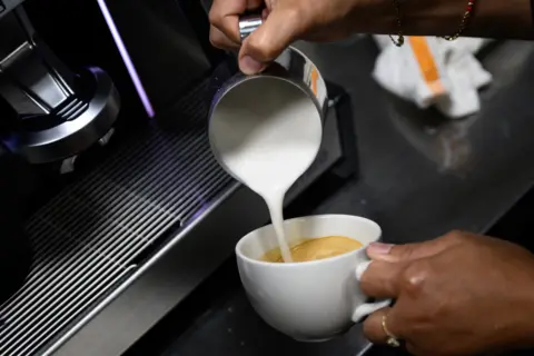 A close-up shot of a barista's hands as they pour milk into a cup of coffee.