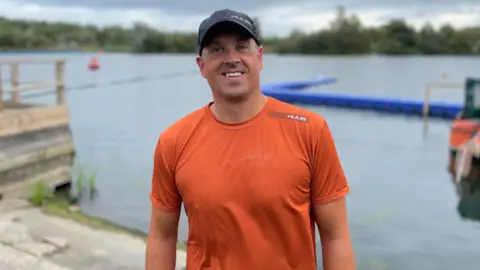 Man in dark blue baseball cap and orange t-shirt in front of a lake