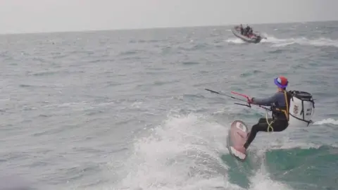 Pain de Minuit - Andoni Martin Marescaux kitesurfing on the sea with a large white barrel on his back. There is a small RIB boat in the background.