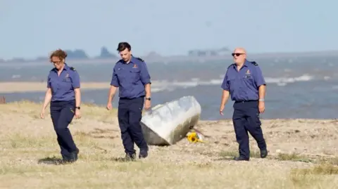Three people wearing navy shirts and trousers walking in a line along the sand. The sea is behind them and there is a bouquet of yellow sunflowers on the sand.