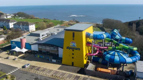 North Yorkshire Council A yellow building with a logo reading Alpamare on the side. Several brightly coloured tube waterslides are exiting the side of the building and weaving around each other with the sea in the distance.