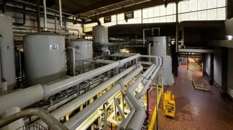 Grey factory machinery with pipes and building materials in front of factory windows