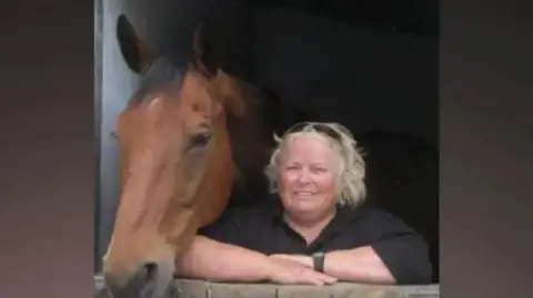 Family photo Wendy Buckney looking out from a stable door alongside a horse. She has blonde hair and wears a black top and her arms are folded, resting on the stable door