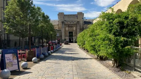 A long stretch down to a hotel which is in the building of a former prison.There are green trees on wither side and a blue sky ahead.
