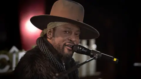 Shahar Azran/Getty Images D'Angelo is seen with a brow hat, furry jacket and scarf. He is in profile and looking into the camera, smiling. He is singing into a mounted mic.