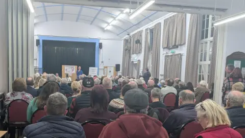Rows of people sat crammed into a community hall, with a man speaking at the front