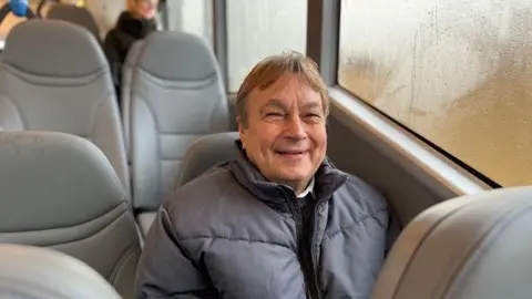 Mr Amess is smiling while sitting in a grey bus seat. He is wearing a grey jacket.