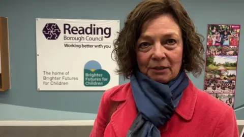 Prof Julia Waters wearing a red coat and navy blue scarf, standing in front of a Reading Borough Council logo sign 