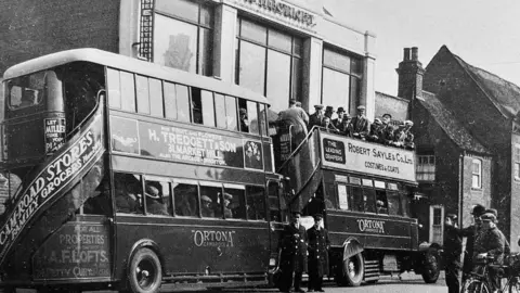 Royston Town Football Club Hop on and hop off buses carry supporters from Royston for their Creake Shield Semi final at Histon in 1931. The vehicles are full with fans wearing flat caps. 