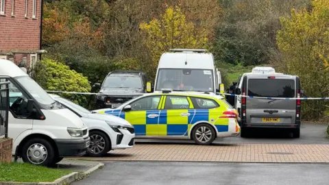 BBC A marked police car parked next to five other vehicles. Police tape can be seen across the end of a quiet cul-de-sac. Beyond the end of the road is a field and some trees. 