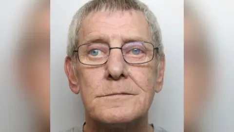 A mugshot of an elderly man with short grey hair and glasses. He is wearing a grey top.