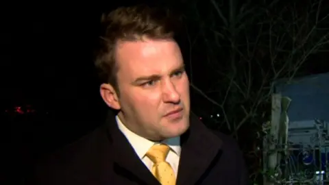 A still of John McGahon. He is standing in the dark in front of a tree. He is wearing a dark jacket, a white collared shirt and a yellow tie. He is looking to a reporter on the other side of the camera.